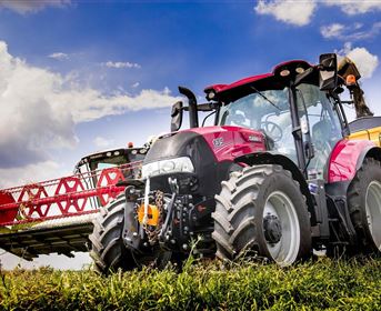 Ein roter Traktor steht auf einem Feld, neben ihm ist eine Erntemaschine zu sehen. Der Himmel ist blau mit einigen Wolken.