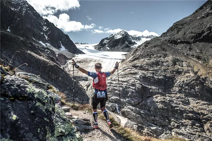 Ein Wanderer steht triumphierend auf einem Bergpfad und genießt die Aussicht auf schneebedeckte Gipfel. Im Hintergrund ist ein Gletscher und ein klarer Himmel zu sehen.