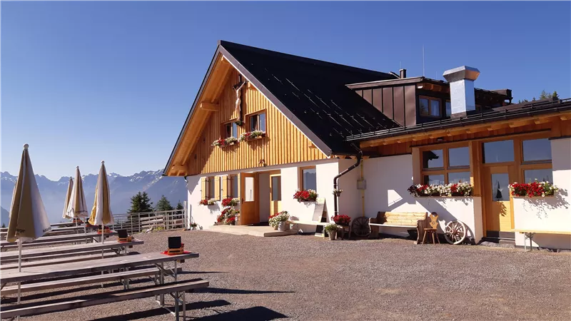 A cozy alpine house with a wooden facade and colorful flowers. In the foreground, there are empty benches and sun umbrellas, with mountains in the background.