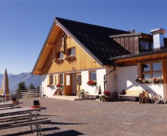 Ein gemütliches alpenländisches Haus mit Holzfassade und bunten Blumen. Im Vordergrund sind leere Sitzbänke und Sonnenschirme zu sehen, mit Bergen im Hintergrund.
