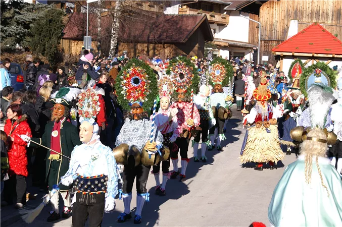 Eine bunte Festparade mit Menschen in traditionellen Kostümen und aufwändigen Kopfschmuck. Im Hintergrund sind Zuschauer und typische alpine Architektur zu sehen.