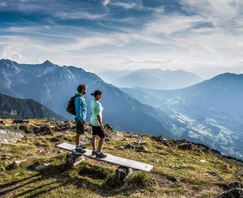 Ein Paar steht auf einer Bank und blickt auf eine malerische Berglandschaft. Die vielen Berge und das weite Tal sind unter einem klaren Himmel zu sehen.