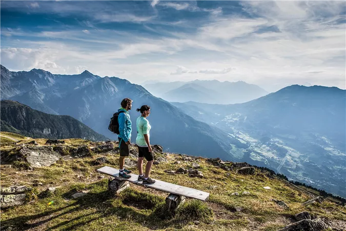 A couple stands on a bench and gazes at a picturesque mountain landscape. The many mountains and the wide valley can be seen beneath a clear sky.