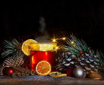 Ein festlicher Drink in einer Tasse, verziert mit einer Orangenscheibe. Umgeben von Tannenzweigen, Zimtstangen und Weihnachtsdekoration.