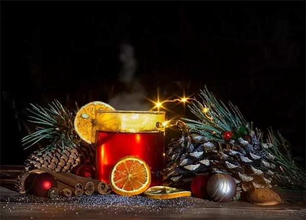 Ein festlicher Drink in einer Tasse, verziert mit einer Orangenscheibe. Umgeben von Tannenzweigen, Zimtstangen und Weihnachtsdekoration.