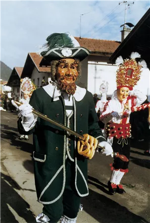Eine festlich gekleidete Person mit einer Maskerade und einem Schlüssel in der Hand. Im Hintergrund sind weitere Teilnehmer der Feierlichkeiten zu sehen.