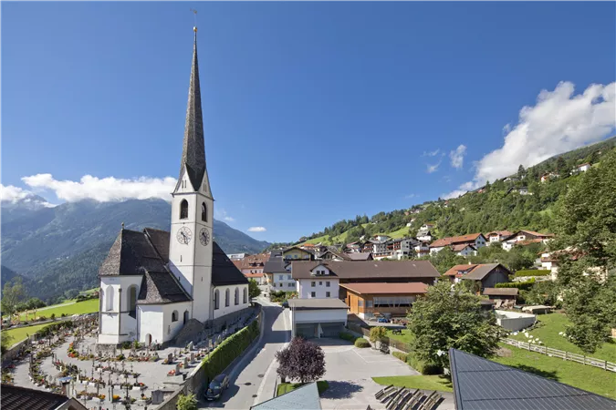 Eine malerische Kirche mit spitzem Turm steht im Vordergrund. Im Hintergrund sind grüne Hügel und traditionelle Häuser zu sehen.