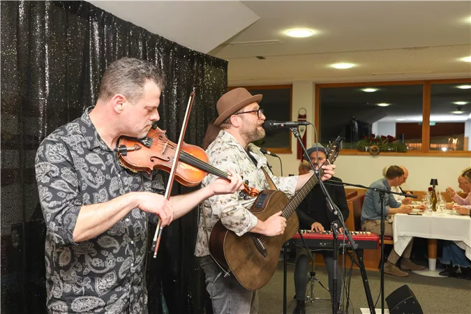 A live band is playing in a cozy room. Two musicians are in the foreground, one with a violin and the other with a guitar.