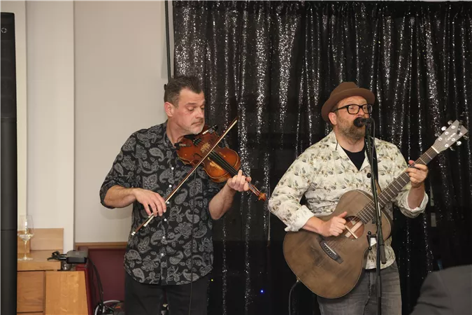 Two musicians are performing on a stage. The man on the left plays the violin, while the man on the right plays the guitar and sings.