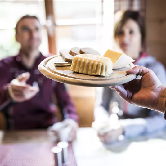 Eine Hand hält einen Teller mit verschiedenen Käsesorten. Im Hintergrund sitzen zwei Personen und schauen interessiert auf das Essen.