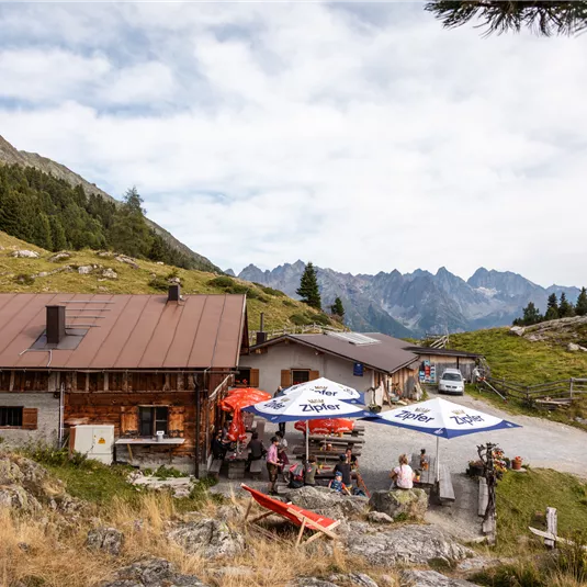 Eine rustikale Hütte in den Bergen mit mehreren Menschen im Freien. Die Umgebung ist von grünen Wiesen und majestätischen Bergen geprägt.