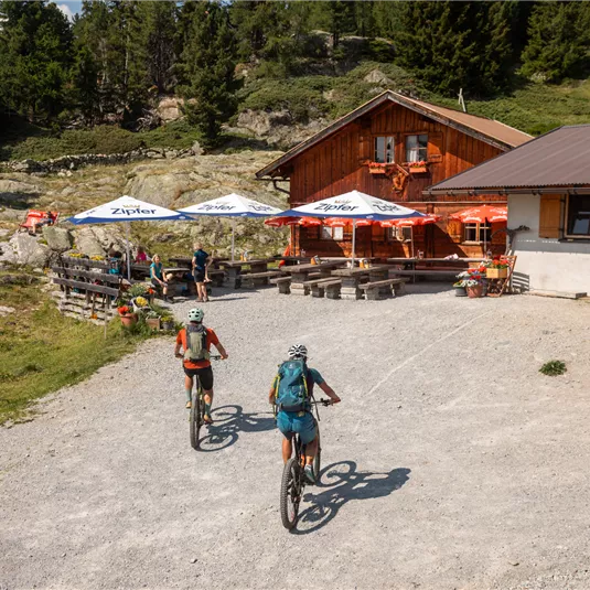 Zwei Radfahrer fahren auf einem Schotterweg zu einer Almhütte. Im Hintergrund sind bewaldete Berge und Sonnenschirme zu sehen.