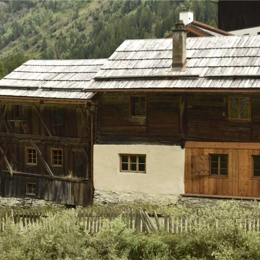 Ein traditionelles Holzhaus in den Bergen, umgeben von grünen Wäldern. Das Gebäude hat ein rustikales Design mit einem steilen Dach und einer einladenden Veranda.