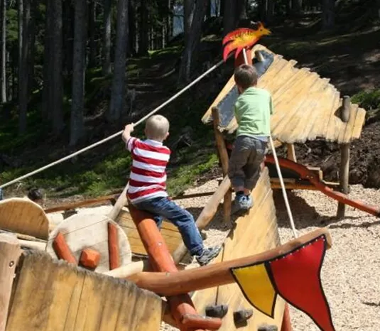 A playground outdoors with wooden elements and a pirate ship. Two children are happily playing and climbing on the play structure.