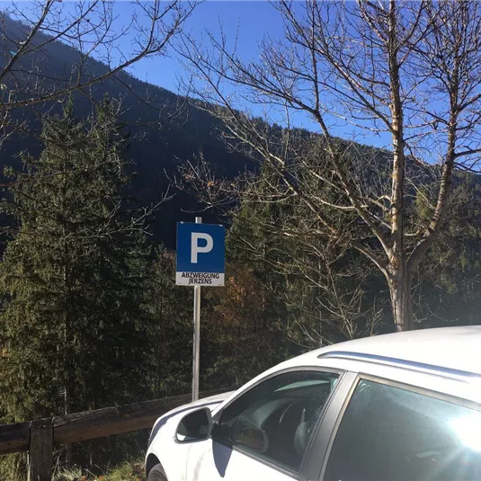 Ein Parkplatzschild steht in der Natur. Im Hintergrund sind Bäume und Berge sichtbar.