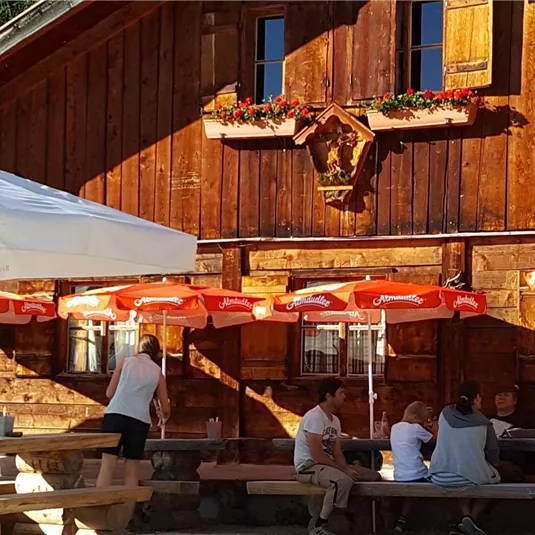 Eine gemütliche Holzstube mit Sonnenschirmen im Freien. Menschen sitzen an Holztischen und genießen die Atmosphäre.