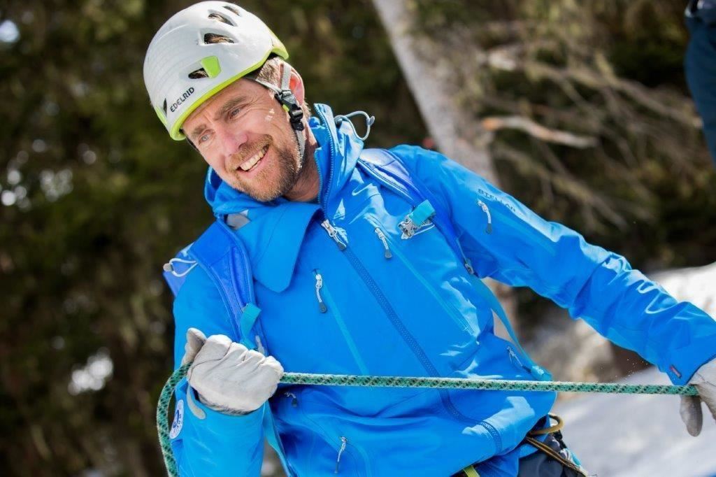 Ein kletternder Mann in blauer Winterkleidung lächelt während einer Bergbesteigung. Er trägt einen Helm und hält ein Seil in der Hand.