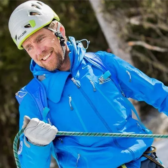Ein kletternder Mann in blauer Winterkleidung lächelt während einer Bergbesteigung. Er trägt einen Helm und hält ein Seil in der Hand.