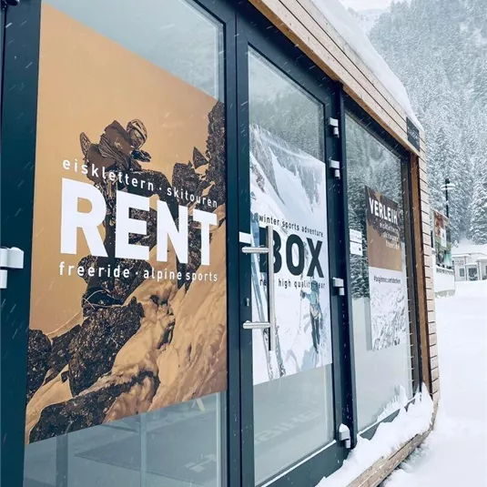 Ein Geschäft für Outdoor-Sportausrüstung mit großen Fenstern. Auf den Schildern steht "RENT BOX" und bietet Vermietung für verschiedene Sportarten an.
