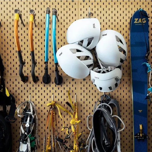 Eine Wand mit Kletter- und Ski-Ausrüstung. Zu sehen sind Helme, Karabiner, Skier und ein Rucksack.
