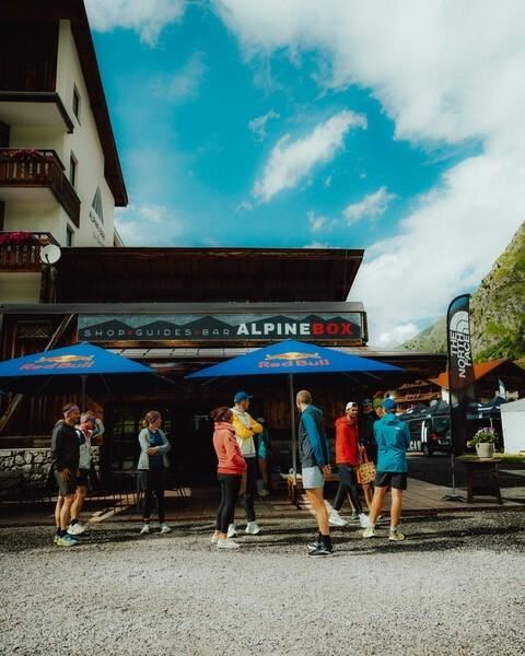 Eine Gruppe von Menschen steht vor einem Geschäft mit dem Namen "ALPINEBOX". Im Hintergrund sind Berge und ein blauer Himmel zu sehen.