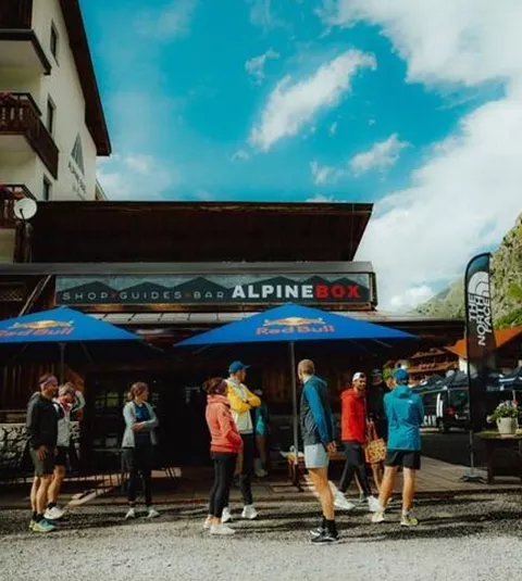 Eine Gruppe von Menschen steht vor einem Geschäft mit dem Namen "ALPINEBOX". Im Hintergrund sind Berge und ein blauer Himmel zu sehen.