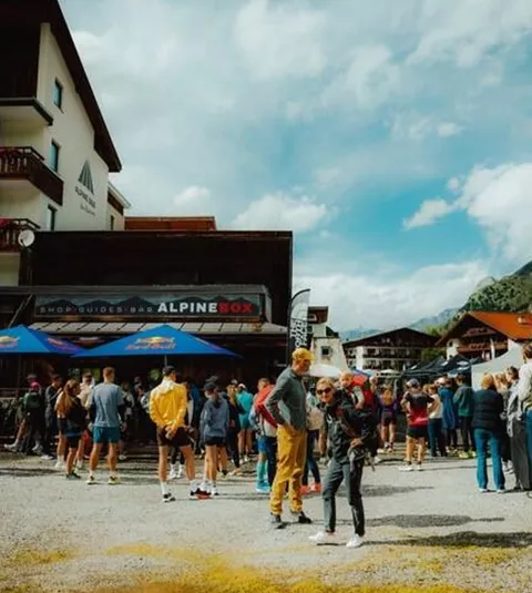 Eine große Menschenmenge versammelt sich vor einem Gebäude in den Bergen. Der Himmel ist bewölkt, und einige Leute tragen Sportkleidung.