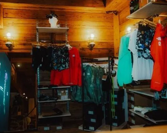 Ein gemütlicher Verkaufsraum mit Holzregalen, die farbenfrohe Kleidung und Accessoires präsentieren. An den Wänden sind stilvolle Lichtakzente zu sehen.