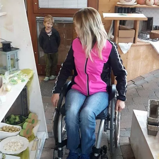Eine Frau im Rollstuhl mit langen blonden Haaren sitzt in einem Geschäft. Ein kleines Kind steht im Hintergrund und schaut neugierig.