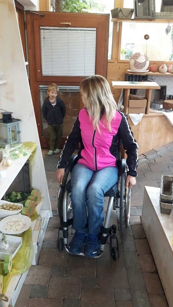 Eine Frau im Rollstuhl mit langen blonden Haaren sitzt in einem Geschäft. Ein kleines Kind steht im Hintergrund und schaut neugierig.