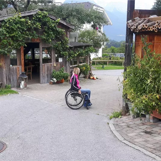 Eine Person im Rollstuhl sitzt an einer Straßenecke in einem ländlichen Bereich. Im Hintergrund sind holzverkleidete Häuser und grüne Pflanzen zu sehen.