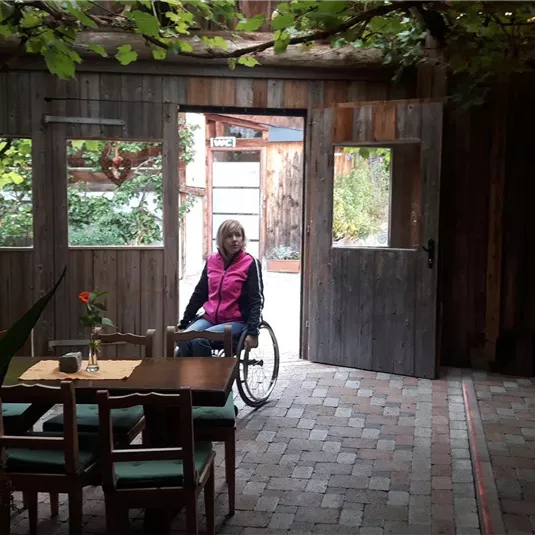 Ein gemütliches Café mit Holzmöbeln und einem offenen Eingang zur Natur. Eine Frau im Rollstuhl sitzt an einem Tisch und genießt die Umgebung.