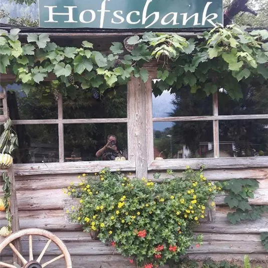 Ein rustikales Gebäude mit einem Schild "Ander's Hofschank" und üppigem Grün. Vor dem Eingang befinden sich dekorative Kürbisse und Blumen.