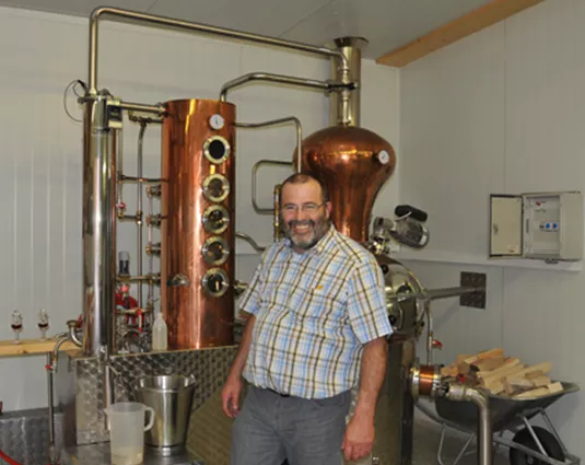 Ein Mann steht neben einer Kupferdampfmaschine in einer Brennerei. Im Hintergrund sind verschiedene Utensilien und Materialien zur Herstellung von Spirituosen zu sehen.