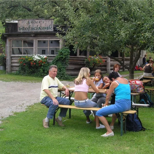 Eine Gruppe von Menschen sitzt an einem Tisch im Freien. Im Hintergrund ist ein Holzgebäude mit Pflanzen zu sehen.