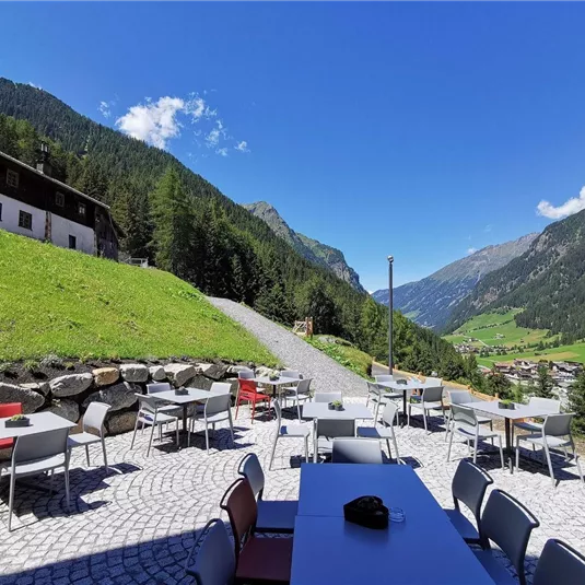 A beautiful outdoor area with tables and chairs, surrounded by a picturesque mountain landscape. The sky is clear and nature is lush green.