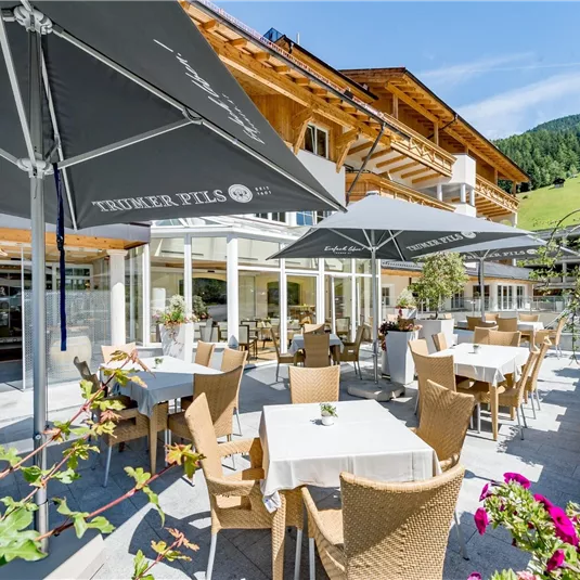 Ein einladendes Café mit sonnigen Terrassen und Schirmen. Im Hintergrund sind grüne Hügel und ein klarer blauer Himmel sichtbar.