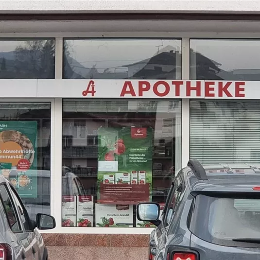 Eine Apotheke mit dem Schild "APOTHEKE" ist zu sehen. Vor der Apotheke stehen mehrere Autos.