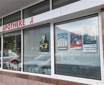 Eine Apotheke mit großen Fenstern und verschiedenen Plakaten. Über dem Eingang steht "APOTHEKE".
