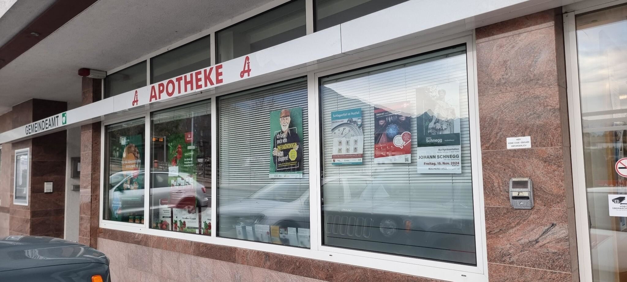 Eine Apotheke mit großen Fenstern und verschiedenen Plakaten. Über dem Eingang steht "APOTHEKE".