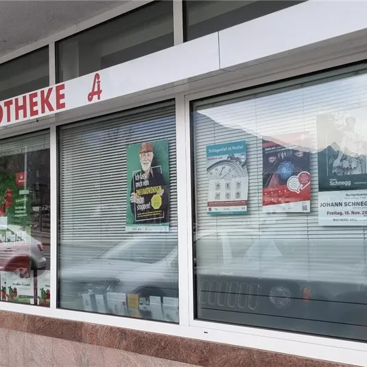 Eine Apotheke mit großen Fenstern und verschiedenen Plakaten. Über dem Eingang steht "APOTHEKE".