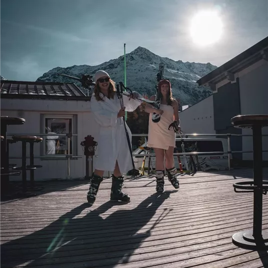 Zwei Frauen in Skikleidung stehen auf einer Holzterrasse. Im Hintergrund sind Berge und die Sonne scheint.