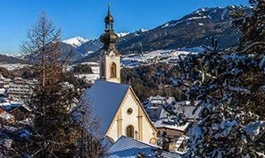 Eine malerische Winterlandschaft mit schneebedeckten Dächern und einer Kirche im Vordergrund. Im Hintergrund erheben sich majestätische Berge unter klarem, blauem Himmel.