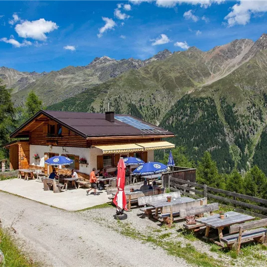 A picturesque mountain cabin surrounded by green forests and majestic mountains. Outside, there are tables and umbrellas, perfect for a break in nature.