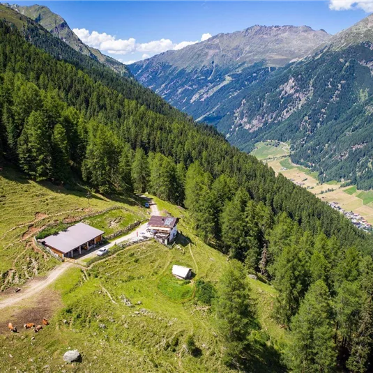 A picturesque mountain landscape with green forests and high mountains in the background. In the middle stands a rustic building, surrounded by natural beauty.