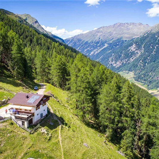 A picturesque mountain cabin surrounded by green forests and tall mountains. The sky is clear and the scenery is breathtaking.