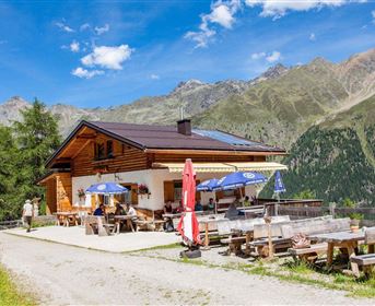 A charming mountain restaurant with a wooden facade and sun loungers. In the background, majestic mountains stretch out under a blue sky.