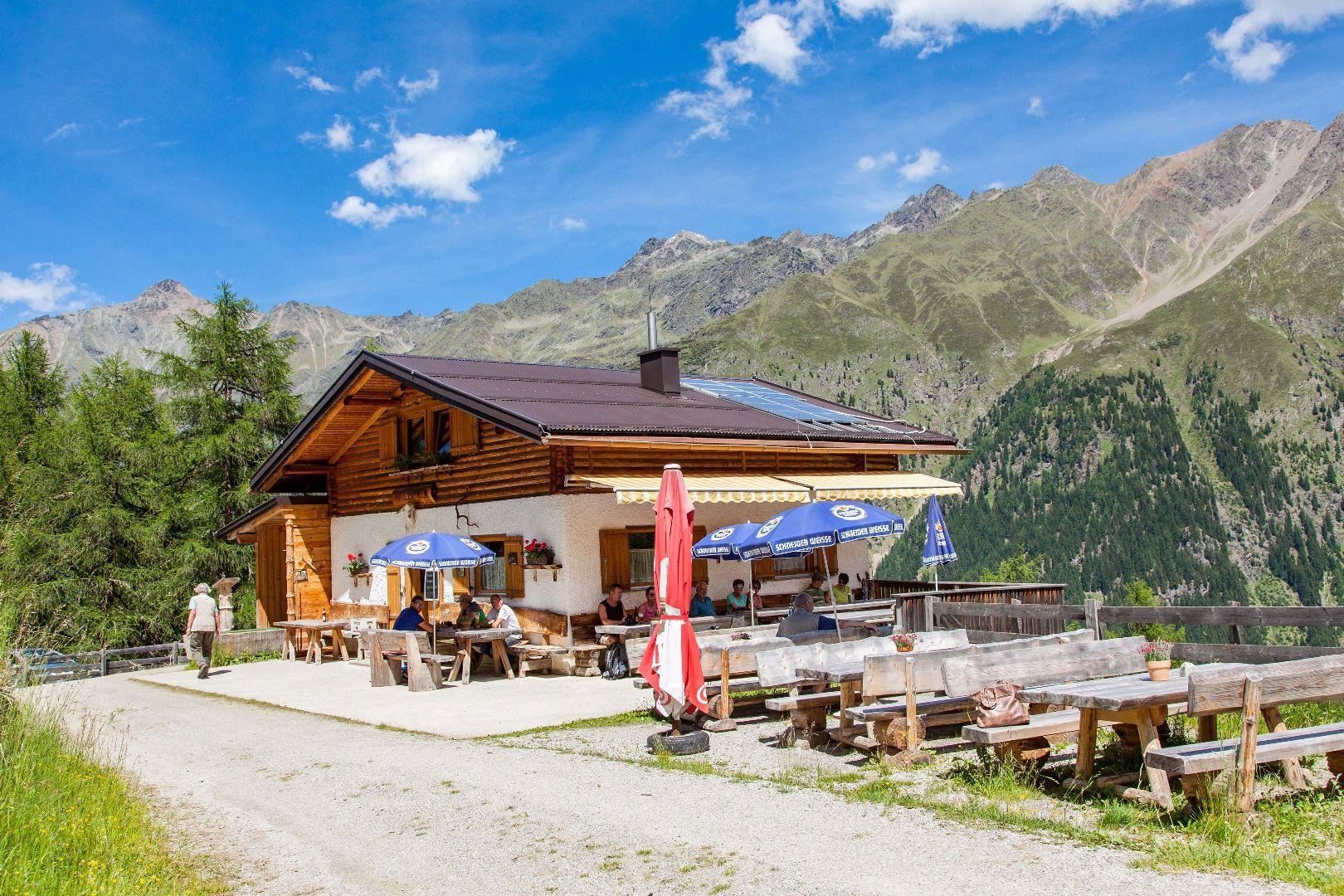 A charming mountain restaurant with a wooden facade and sun loungers. In the background, majestic mountains stretch out under a blue sky.