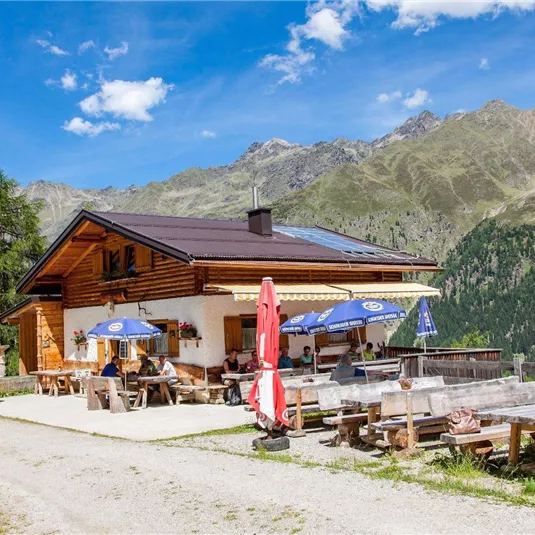 A charming mountain restaurant with a wooden facade and sun loungers. In the background, majestic mountains stretch out under a blue sky.