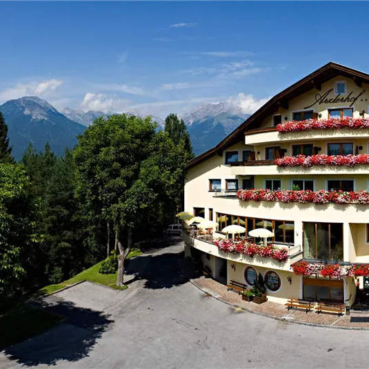 Ein charmantes Hotel mit einem bunten Blumenschmuck an den Balkonen. Im Hintergrund sind grüne Bäume und majestätische Berge zu sehen.